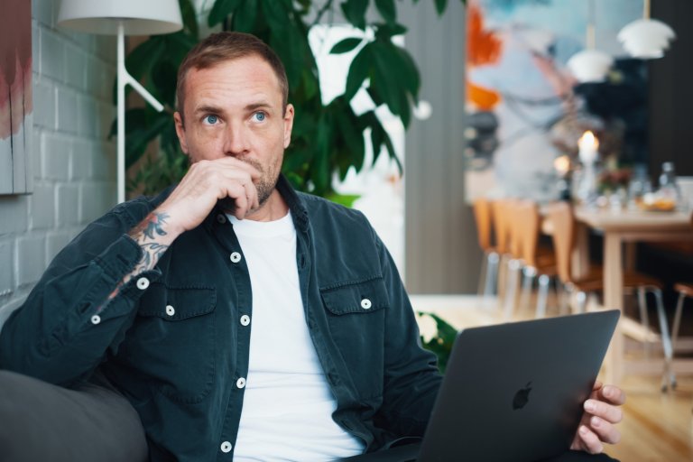 En man sitter på en soffa med en laptop i knät och ser fundersam ut