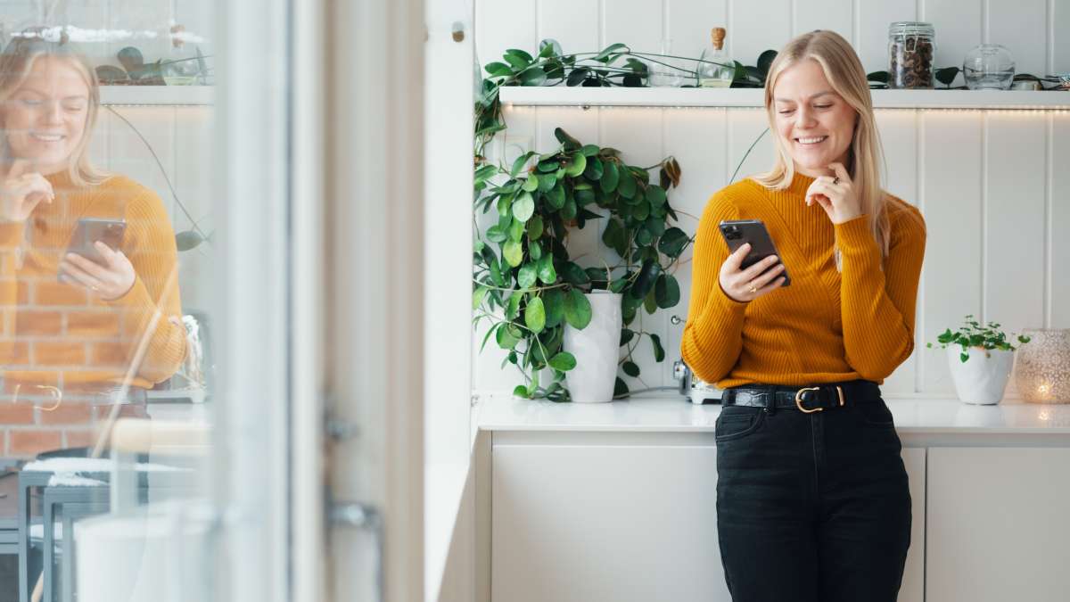 En kvinna står och ler och tittar på en mobiltelefon i handen