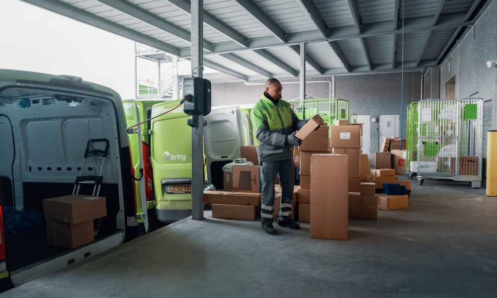 A Bring driver is holding a parcel and stands on a loading dock with several parcels around him.