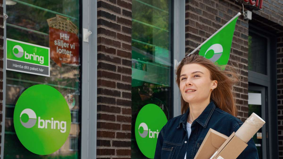 En kvinna har tre paket i handen och står utanför ett ombud med Bring-skyltar.